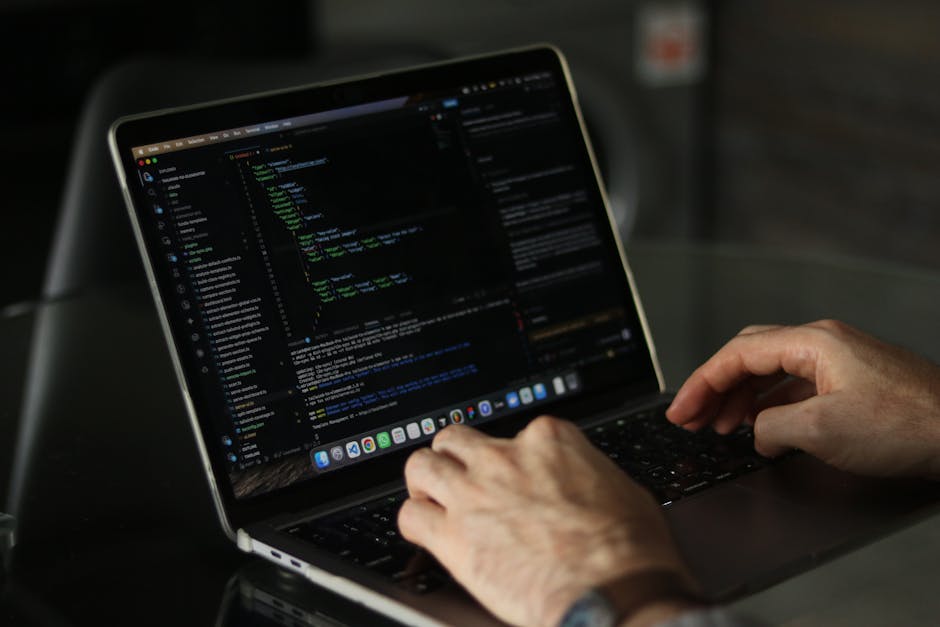 A close-up of a person's hands typing on a laptop keyboard, displaying a dark-themed coding interface with lines of colorful code. The laptop screen shows a text editor with various programming syntax