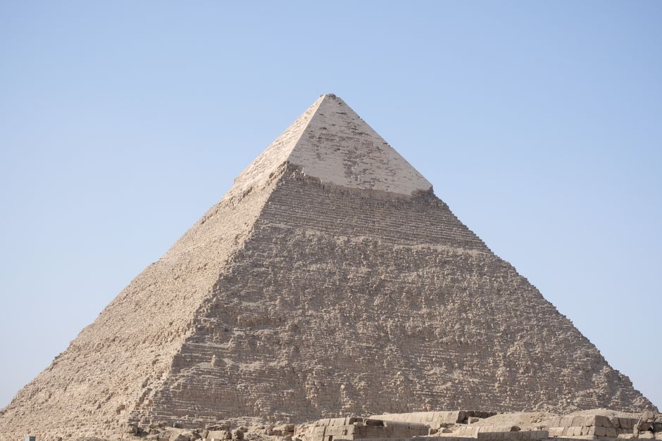A close-up view of the Great Pyramid of Giza showing its weathered limestone blocks arranged in a stepped pattern, with some sections appearing rough and eroded. The pyramid tapers smoothly towards th